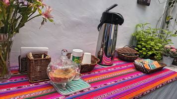 A table with a colorful woven tablecloth, a glass decanter, a thermos, and a basket of books.