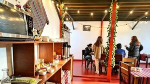 A dining area with wooden tables and chairs, a buffet counter with food and drinks, and a kitchen area with appliances. - Hostal Flor de Loto  (Cusco)