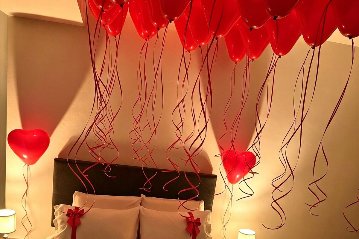 Una habitación de hotel con una cama, almohadas, una botella, vasos y globos rojos.