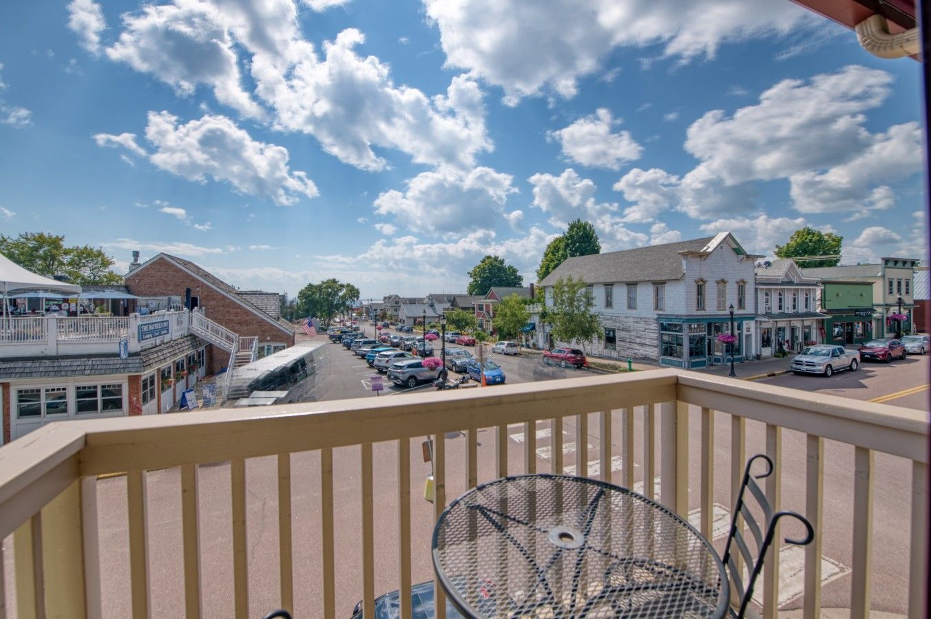 A balcony with a metal table and chairs, overlooking a street with parked cars and buildings.