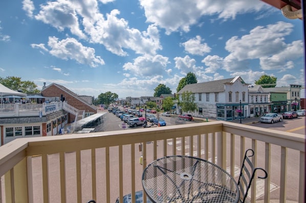 A balcony with a metal table and chairs, overlooking a street with parked cars and buildings.
