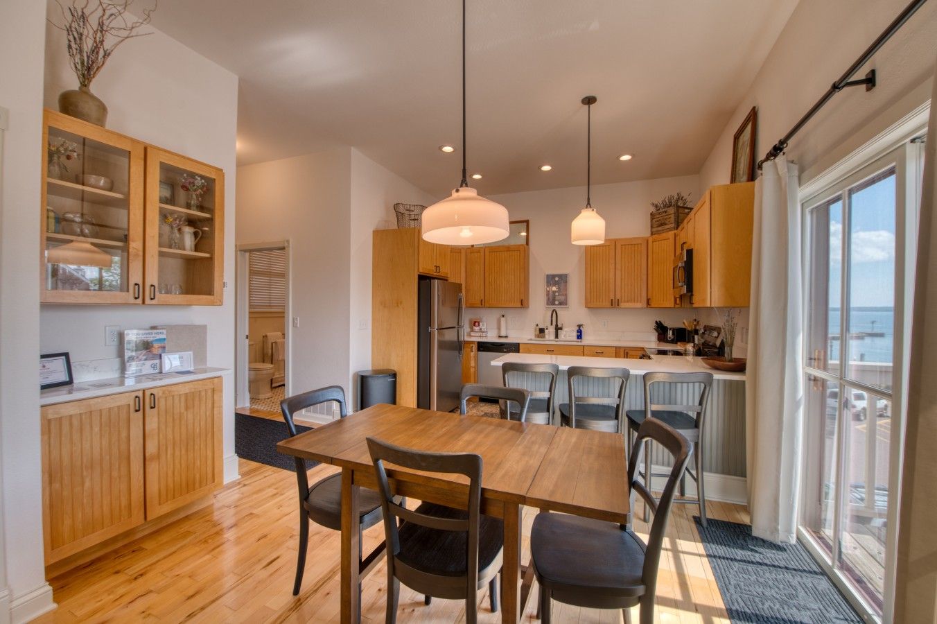 A modern kitchen with wooden cabinets, a dining table with chairs, and pendant lighting.
