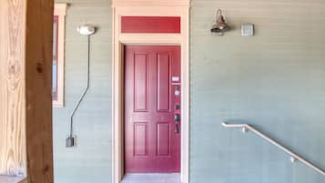 A red door with a keypad lock, a small outdoor light fixture, and a handrail on a wooden porch.