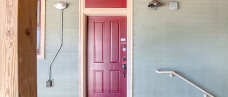 Una puerta roja con cerradura electrónica, una lámpara exterior pequeña y una barandilla en un porche de madera.