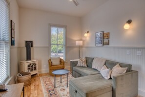 Un salon chaleureux avec un canapé, un fauteuil et une petite table. On y trouve une cheminée, une fenêtre avec des stores et des lampes fixées au mur.