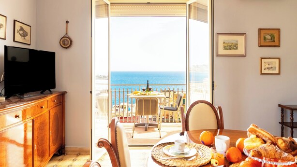 Un balcón con vista al mar, una mesa de comedor con alimentos para el desayuno, un televisor y un armario de madera.