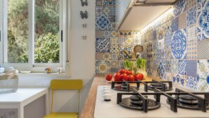 Una cocina con estufa a gas, una silla amarilla, una ventana con vista a la vegetación y un salpicadero de azulejos con diseños azules y blancos.