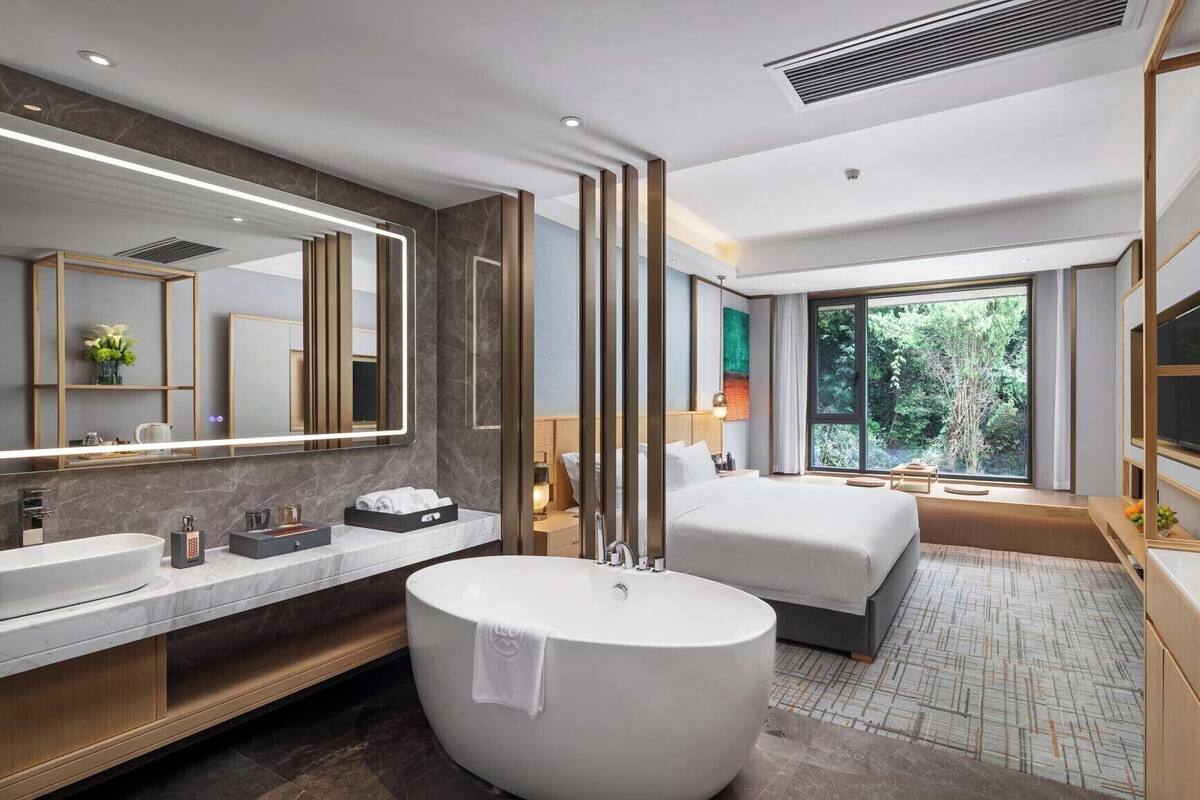 Ein modernes Hotelzimmer mit einer großen Badewanne, einem Doppelbett, einem großen Fenster mit Blick auf Grünflächen und einem Schminktisch mit Spiegel.
