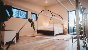 Ein modernes Schlafzimmer mit einem Himmelbett, Holzboden, einem kleinen Nachttisch und einem Fenster mit Blick auf Bäume.