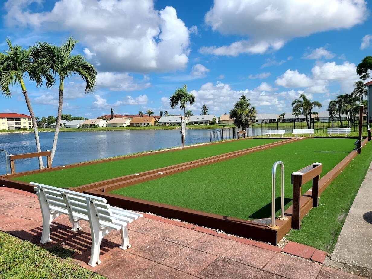 A croquet field with a bench and palm trees, a body of water, and buildings in the background.