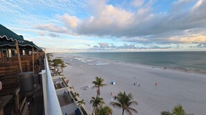 Vista a la playa desde un balcón con terraza de madera, palmeras y una playa de arena.