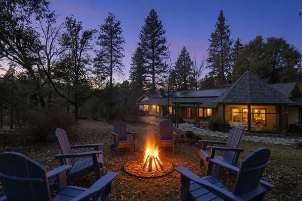 A cozy outdoor seating area with Adirondack chairs around a campfire, a stone fireplace, and a lodge with a covered patio.