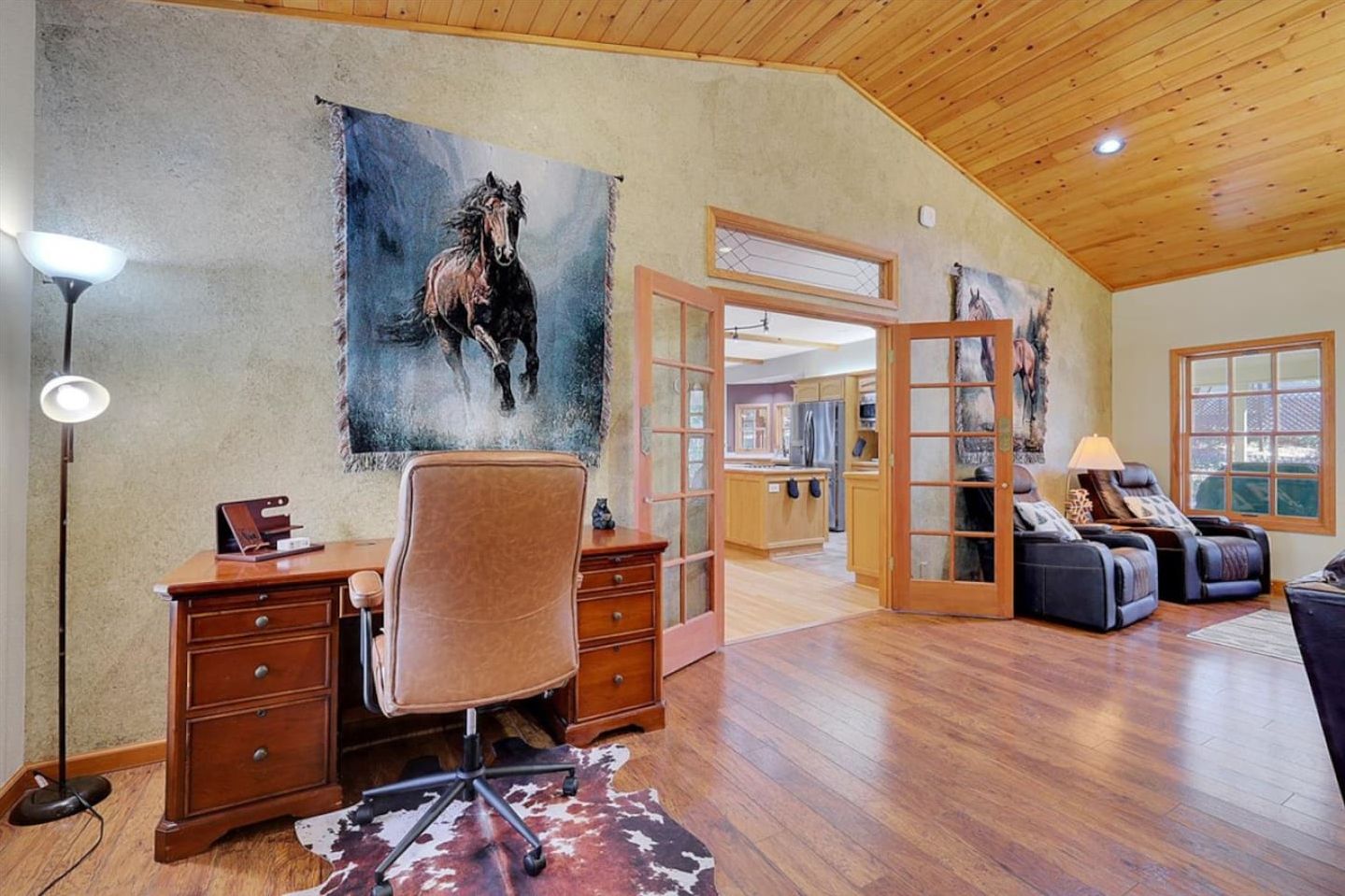 A spacious room with a wooden ceiling, a desk with drawers, a leather chair, a massage chair, and a large painting of a horse on the wall.