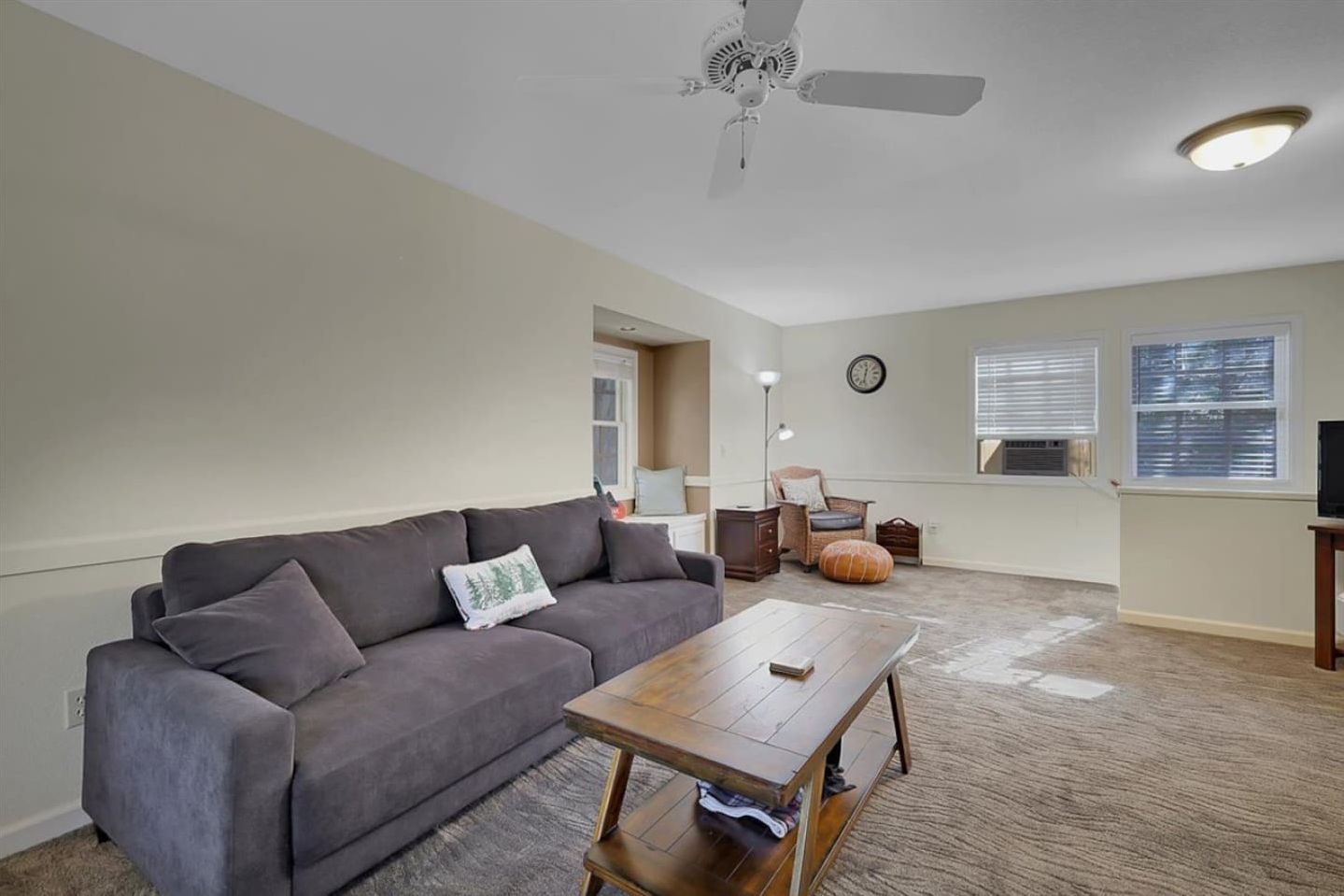 Ein Wohnzimmer mit einem grauen Sofa, einem Holztisch, einem Deckenventilator und einem großen Fenster mit Jalousien.