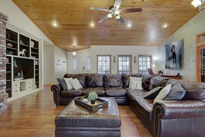 A spacious living room with a large leather sectional sofa, a wooden ceiling with a ceiling fan, and a brick fireplace.