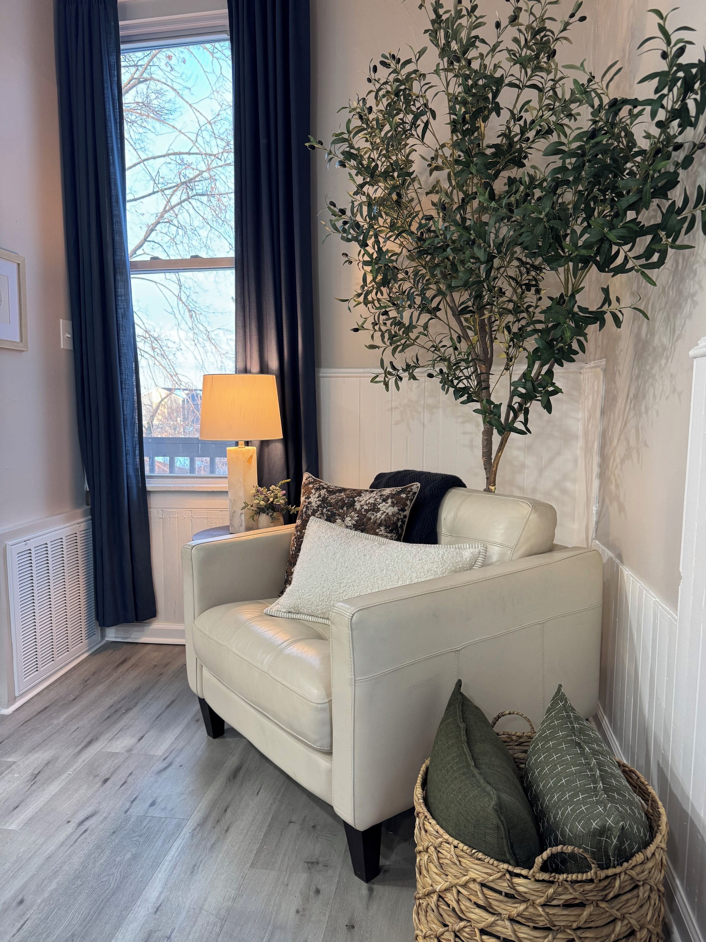 A cozy living room with a beige armchair, a large potted plant, a wicker basket with cushions, and a window with dark curtains.