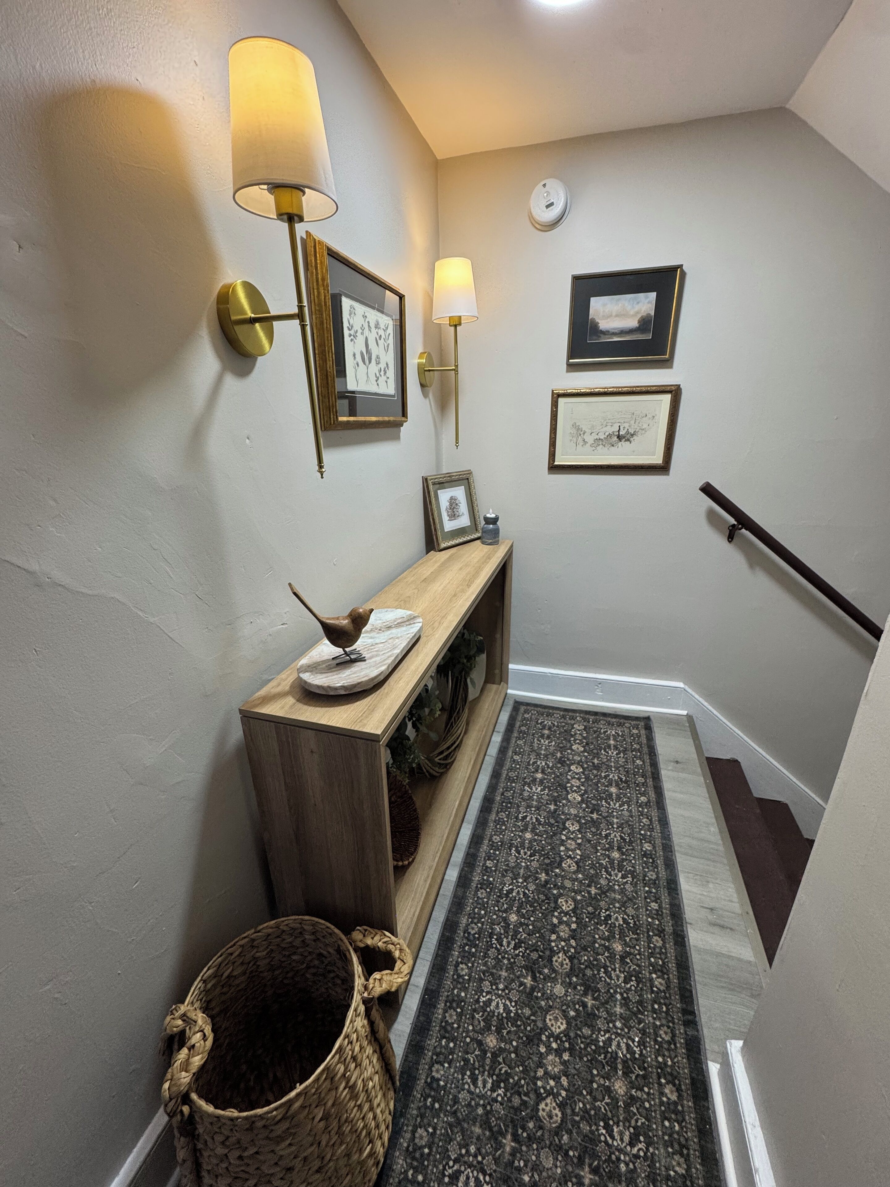 A hallway with a wooden console table, two wall-mounted lamps, framed artwork, a woven basket, and a patterned rug.