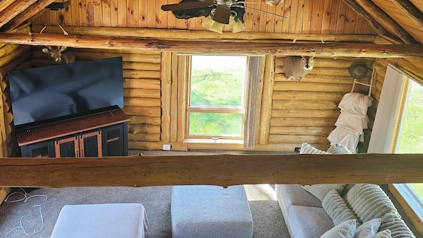 A log cabin interior with a wooden ceiling, a ceiling fan, a mounted television, a sofa with patterned cushions, and a window with a view of the outdoors. - 5-bedroom cabin - Where peaceful vacationing meets nearby adventures  (Wisconsin Dells)