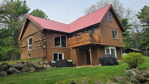 A two-story log cabin with a red roof, a balcony, and a stone wall in the foreground. - 5-bedroom cabin - Where peaceful vacationing meets nearby adventures  (Wisconsin Dells)