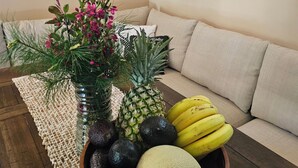 A fruit bowl on a woven placemat, a pineapple, bananas, avocados, and a melon on a wooden table, with a couch and a 'welcome' sign in the background. - 5-bedroom cabin - Where peaceful vacationing meets nearby adventures  (Wisconsin Dells)