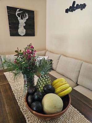 A fruit bowl on a woven placemat, a pineapple, bananas, avocados, and a melon on a wooden table, with a couch and a 'welcome' sign in the background. - 5-bedroom cabin - Where peaceful vacationing meets nearby adventures  (Wisconsin Dells)