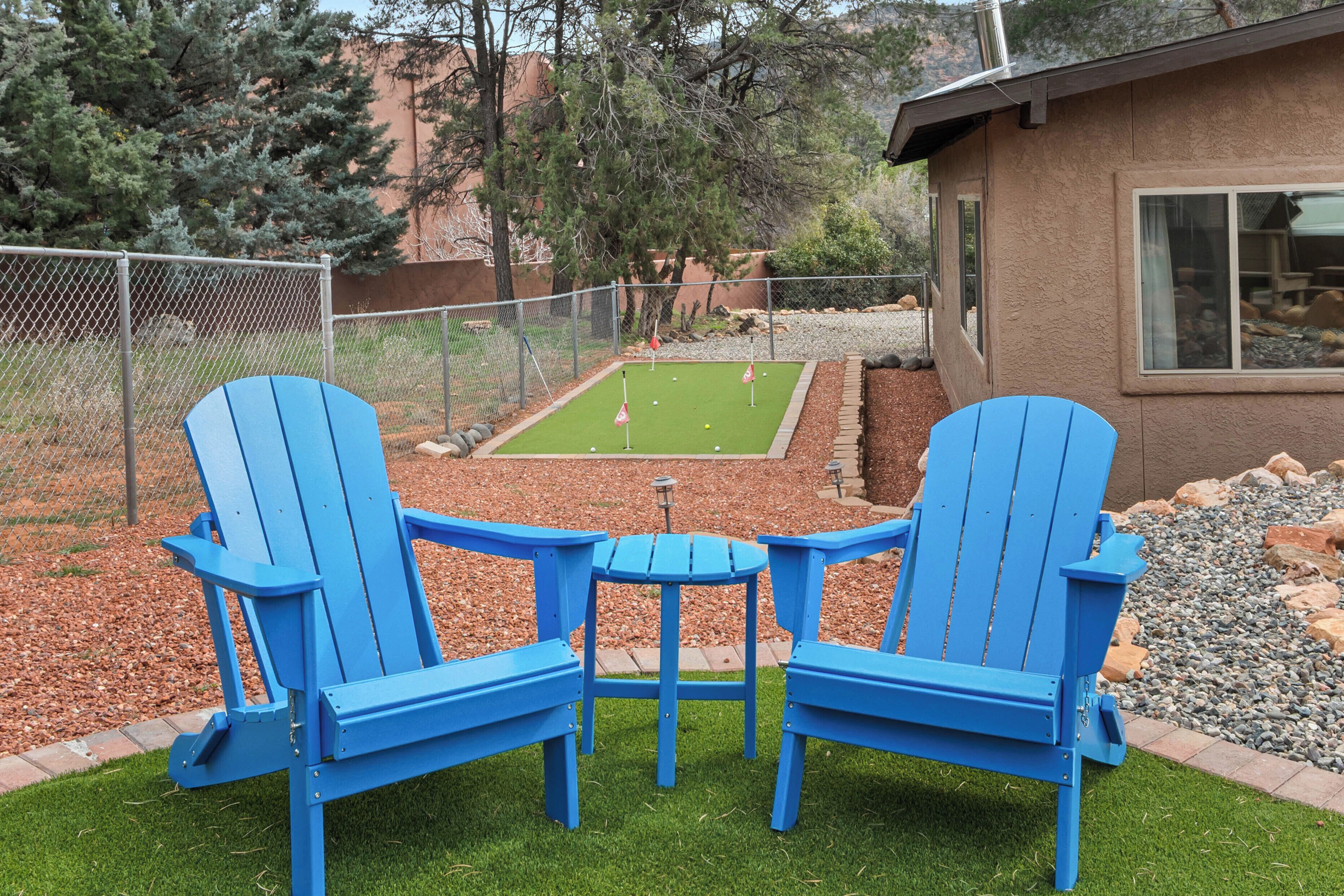 Un patio trasero con dos sillas Adirondack azules, una mesita y un campo de práctica de golf.