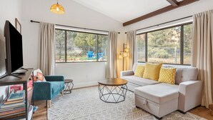 Ein modernes Wohnzimmer mit Sofa, Couchtisch und Fernseher. Große Fenster mit Vorhängen, ein Bücherregal und ein gemustertes Teppich sind ebenfalls vorhanden.