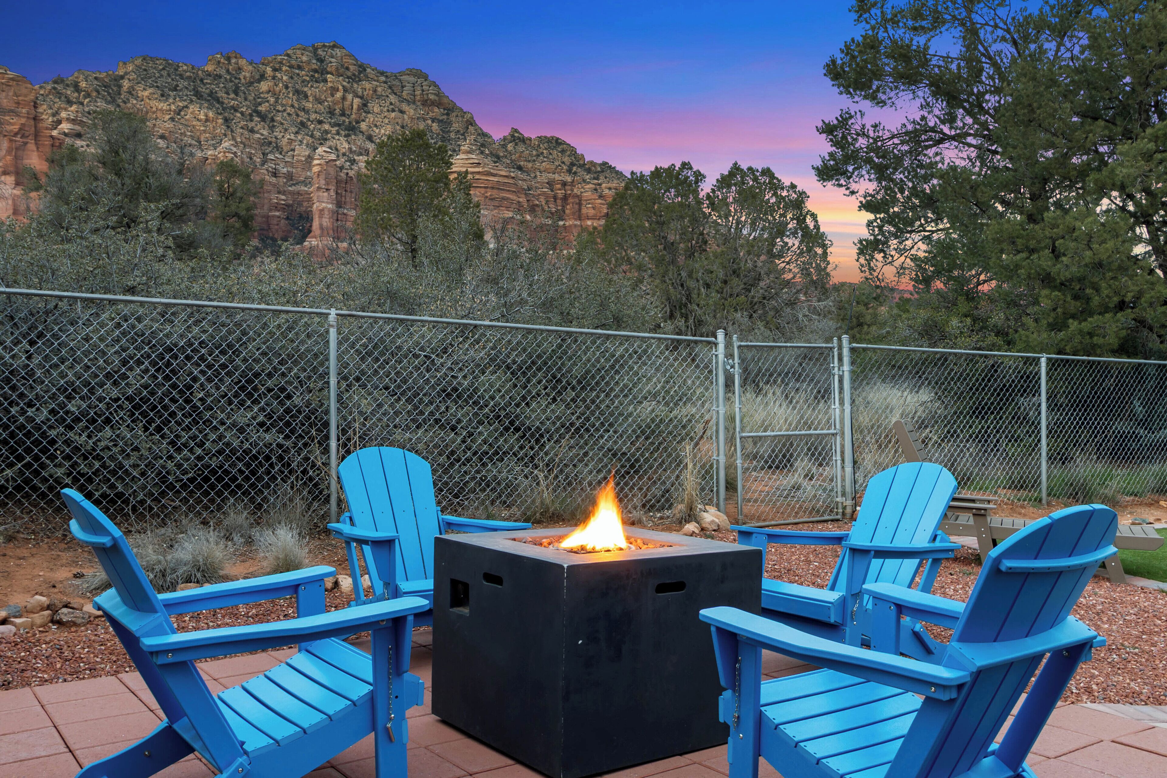 A square fire pit with a fire, surrounded by blue Adirondack chairs, set against a backdrop of red rock formations and a twilight sky.