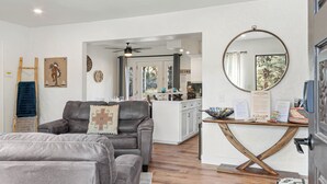 Ein modernes Wohnzimmer mit einem grauen Ledersofa, einem Holztisch und einem großen Spiegel. Im Hintergrund ist eine Küche mit weißen Schränken und einem Essbereich zu sehen.