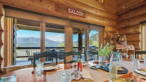 Un intérieur en bois, avec une enseigne indiquant « SALOON DE COWBOYS », des tables à manger dressées avec des assiettes, des verres et un pichet, et une vue sur les montagnes par de grandes fenêtres.