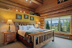Ein schlicht eingerichtetes Schlafzimmer im Holzblockhaus-Stil mit einem großen Bett, einem Holz-Kopfende und Blick ins Freie durch große Fenster.