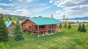 Une grande cabane en rondins avec un toit vert, une terrasse et une vue sur un lac et des montagnes.