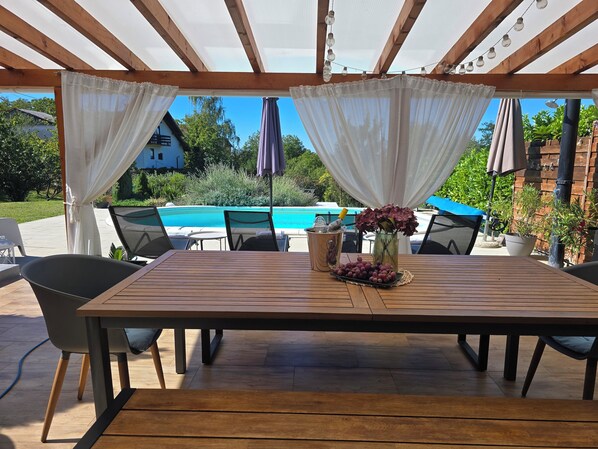 Une terrasse extérieure avec une table en bois, des chaises, une piscine et une pergola décorée de guirlandes lumineuses.