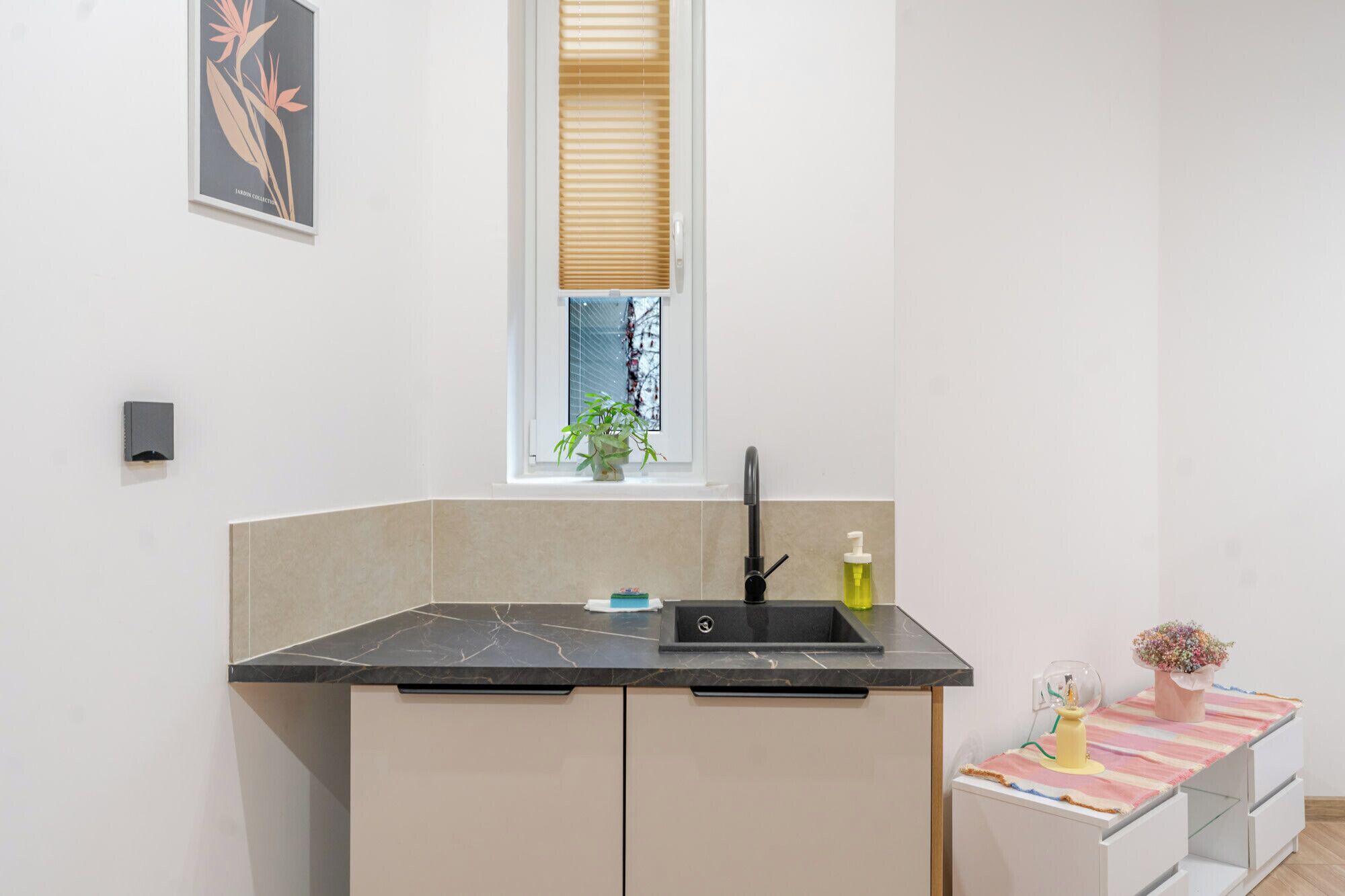 Une cuisine compacte avec un plan de travail noir, un évier, une fenêtre avec des stores et une petite plante en pot.