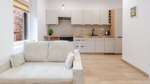 Eine moderne Küche mit weißen Schränken, einer Holz Arbeitsplatte und einem eingebauten Backofen. Ein Fenster mit Jalousien, eine Pflanze auf der Arbeitsplatte und ein weißes Sofa im Vordergrund.