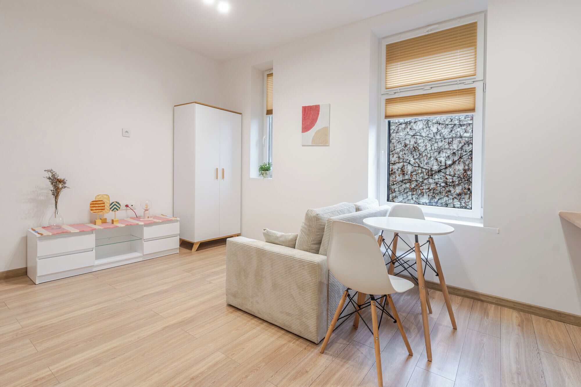Een moderne woonkamer met een bank, een eettafel met stoelen, een witte kledingkast en een raam met jaloezieën.