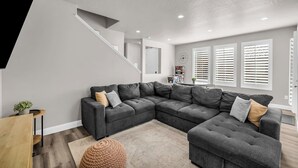 Un salon moderne avec un canapé d’angle gris, une table basse en bois et un pouf rond en fibres tressées.