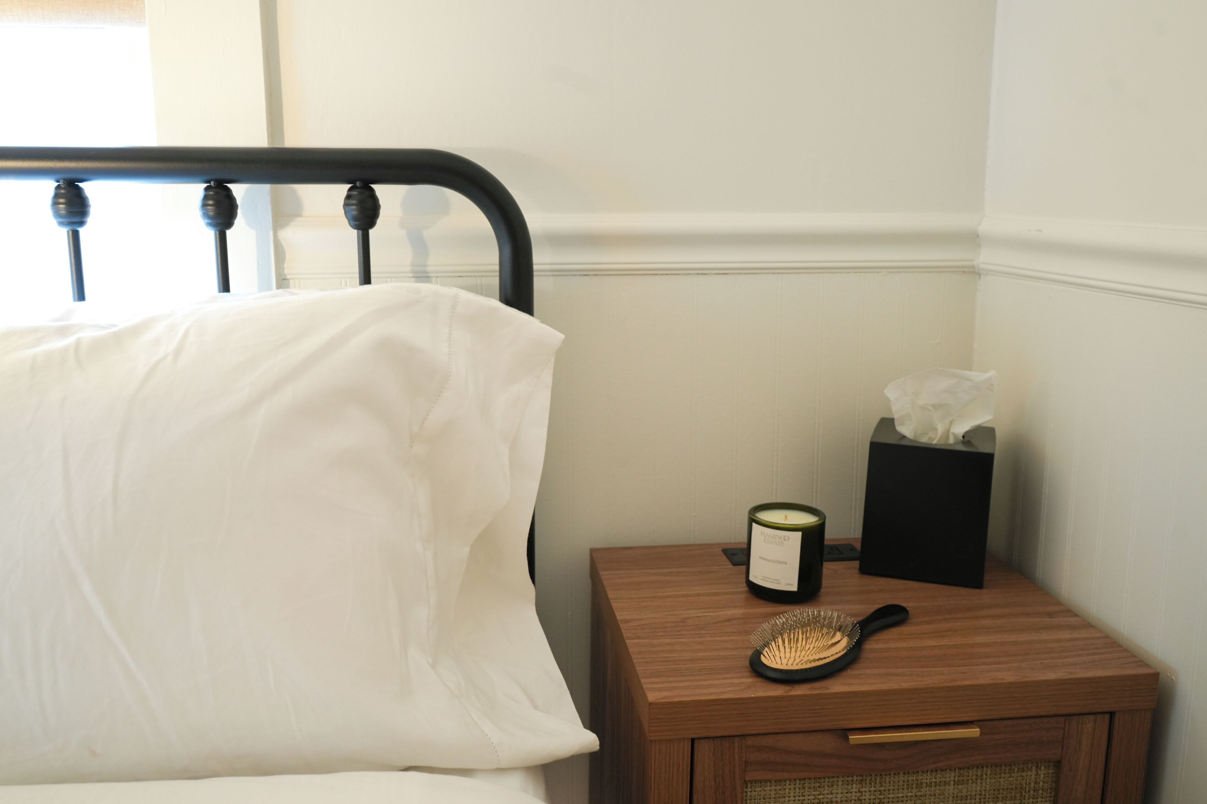 A neatly made bed with white linens, a bedside table with a candle, a tissue box, and a hairbrush.