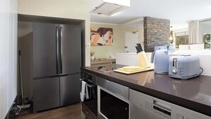 Eine moderne Küche mit Edelstahl-Kühlschrank, Backofen und Geschirrspüler. Eine Arbeitsplatte mit Toaster und einem Messerblock ist vorhanden. Im Hintergrund sind eine Steinwand und ein Fenster mit Vorhängen zu sehen.