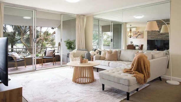 Ein modernes Wohnzimmer mit einem weißen Sofa, einem runden Couchtisch und einem Fernseher auf einem Holzgestell. Große Fenster bieten einen Ausblick ins Freie.