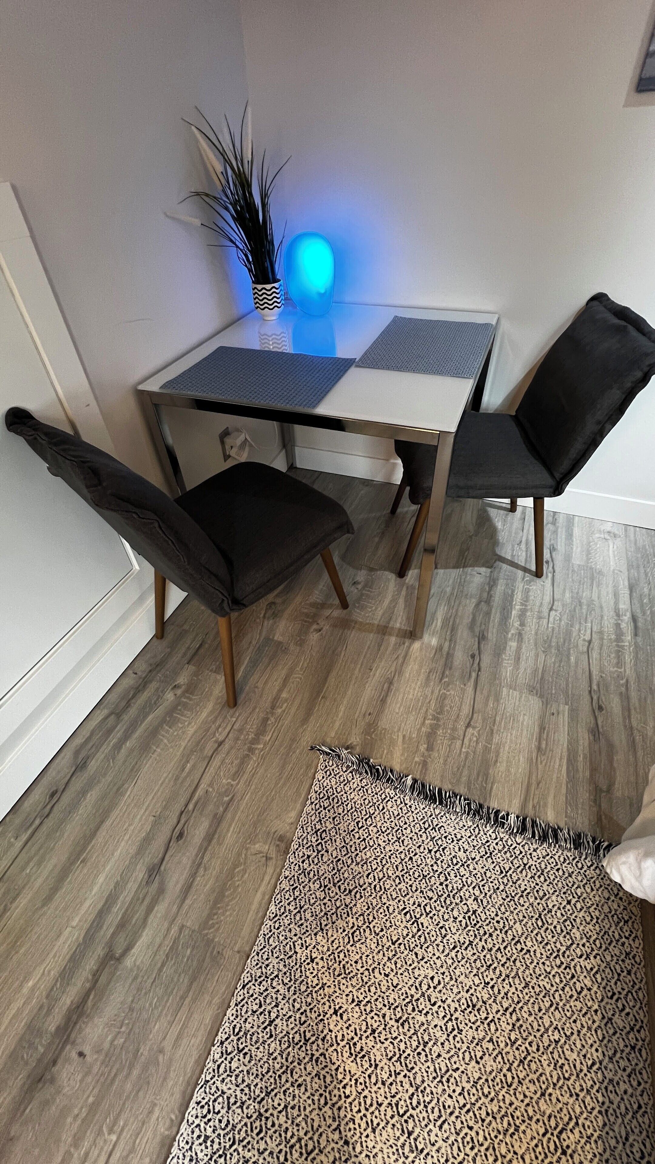 A dining area with a table, chairs, a rug, and a blue-lit object on the wall.
