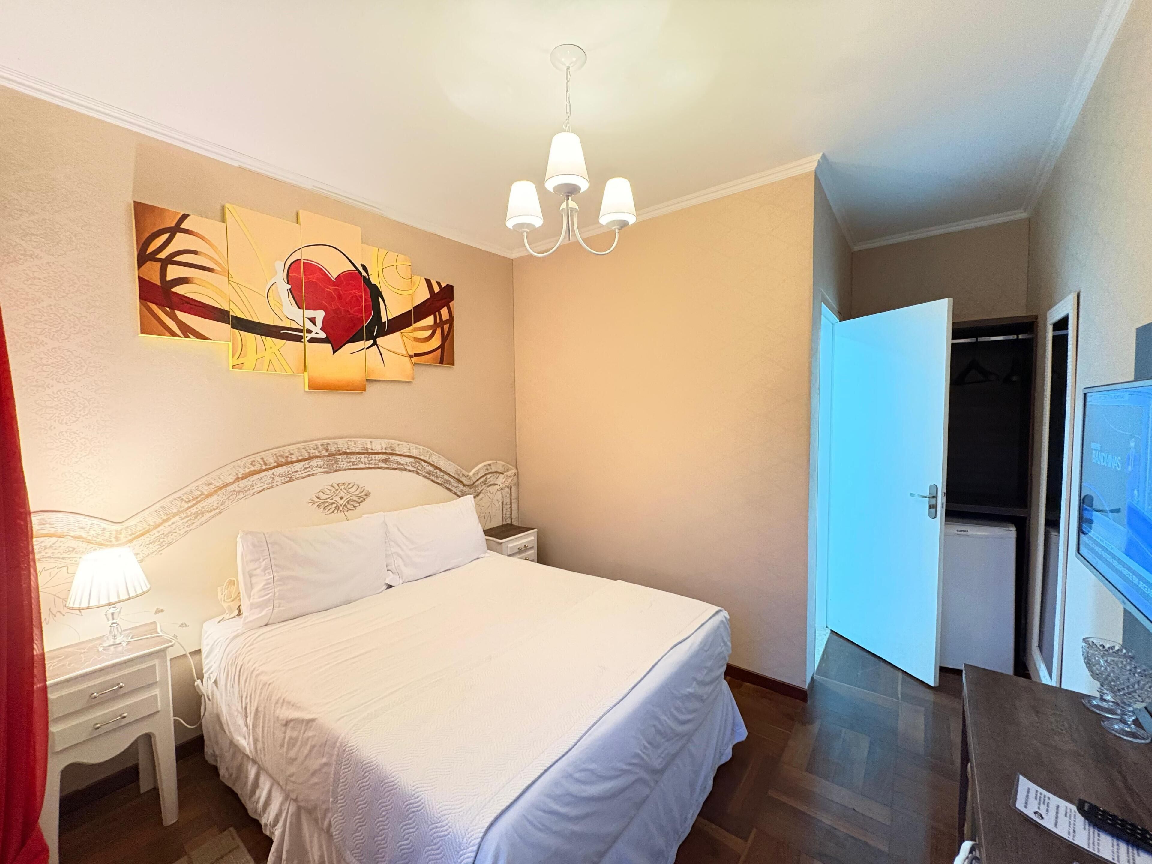 Un dormitorio con una cama grande, una mesita de noche, una lámpara, un cuadro en la pared, una araña y un televisor sobre un mueble.