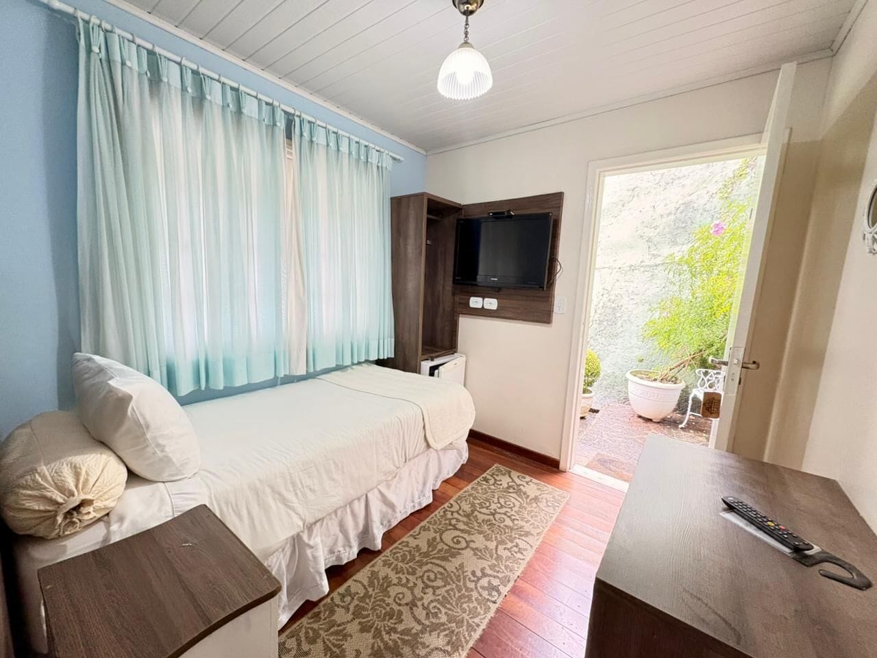 A bedroom with a bed, a TV mounted on the wall, a small table, and a door leading to a balcony with potted plants.