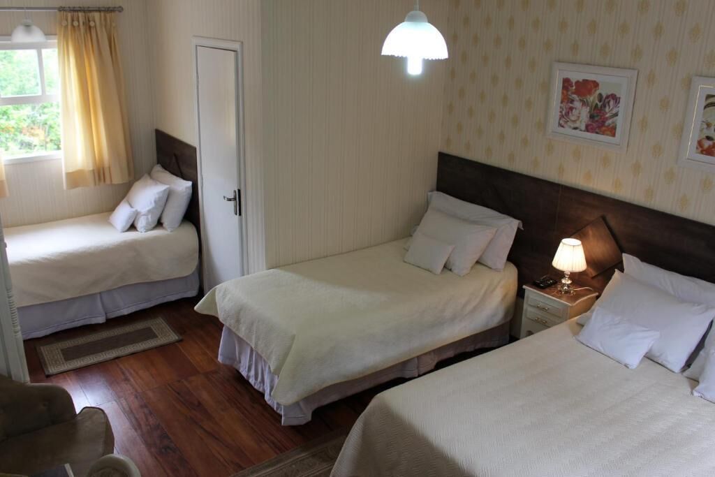 A hotel room with two single beds, a nightstand with a lamp, a wooden floor, and a window with curtains.