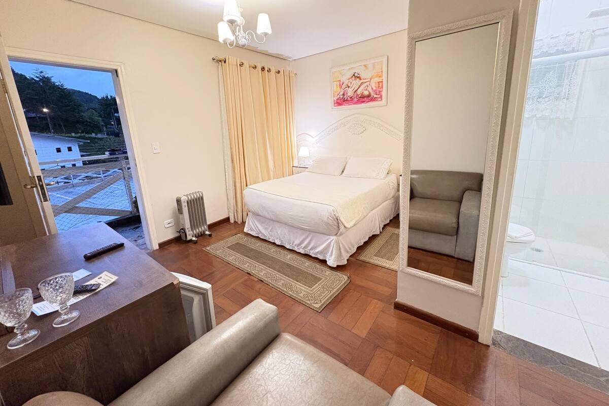 A bedroom with a bed, a chair, a radiator, a desk with a chair, and a view of the outside.