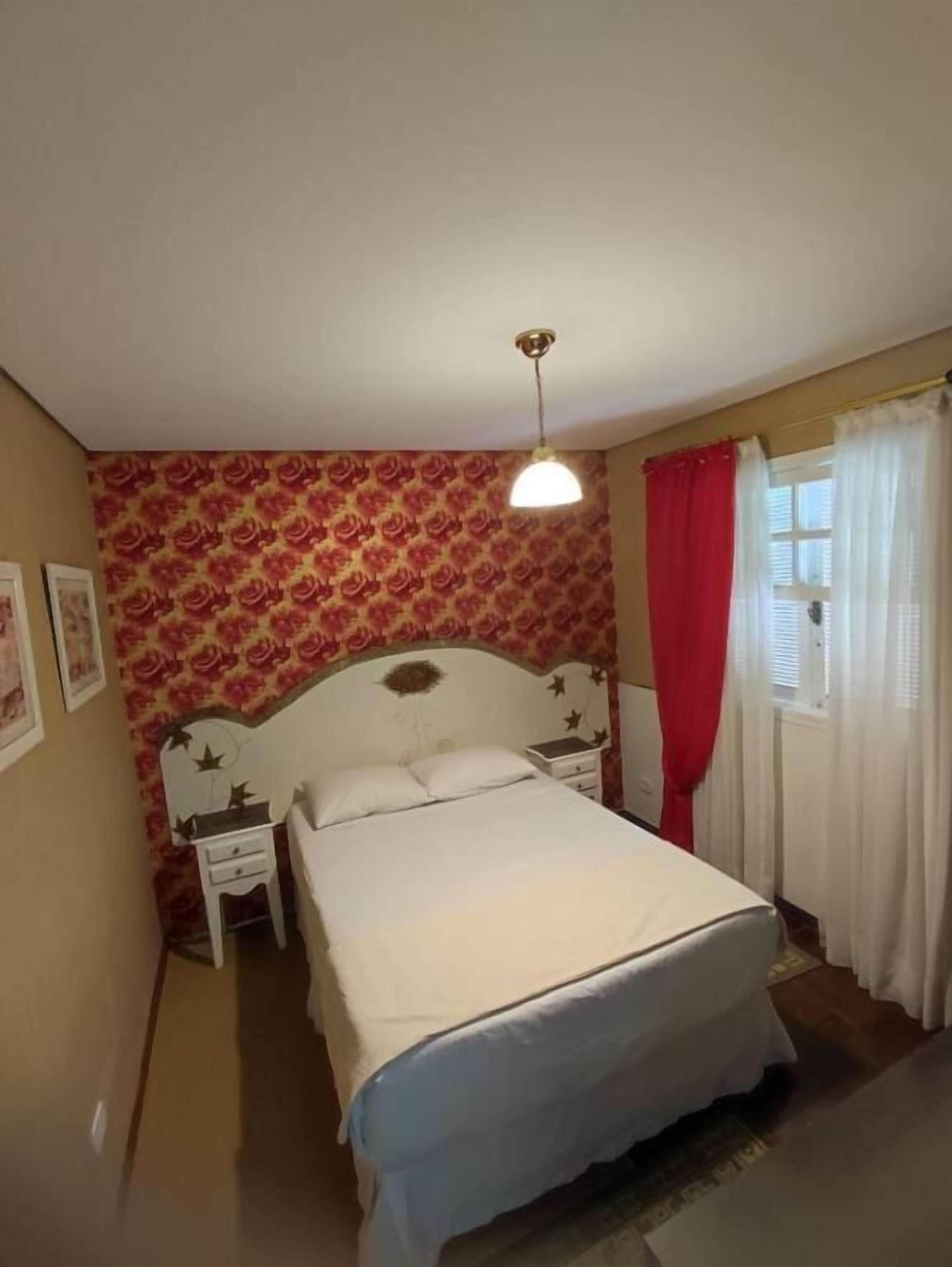 A bedroom with a bed, a nightstand, a wall clock, and a window with curtains.