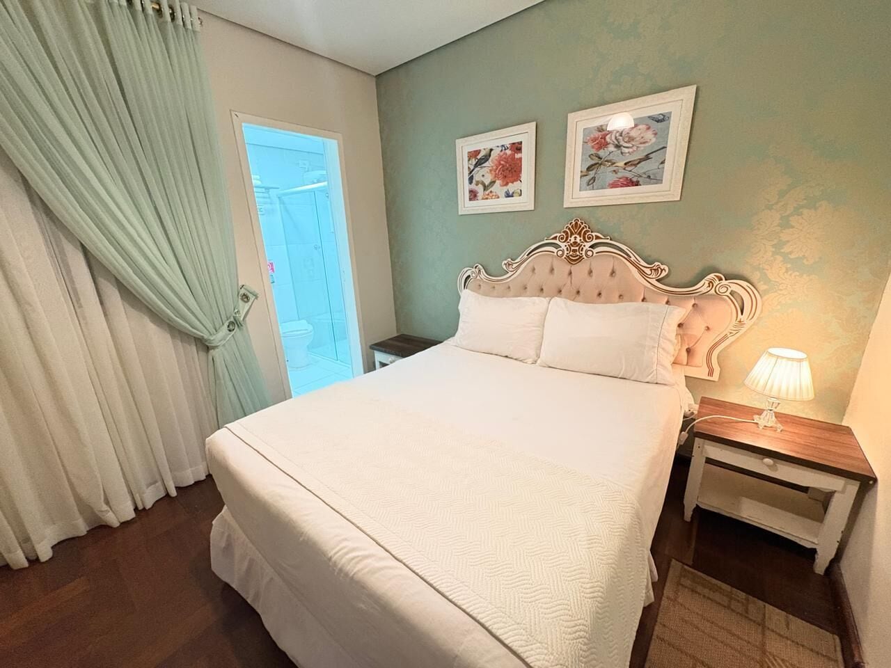 A bedroom with a large bed, a nightstand with a lamp, and two floral wall art pieces.