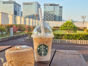 Ein Starbucks-Becher mit Stroh auf einem Holztisch, ein Gebäckstück und eine Stadtlandschaft im Hintergrund.