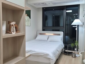 Quarto de hotel moderno com cama, prateleira de madeira com um objeto decorativo, janela e luminária de chão.