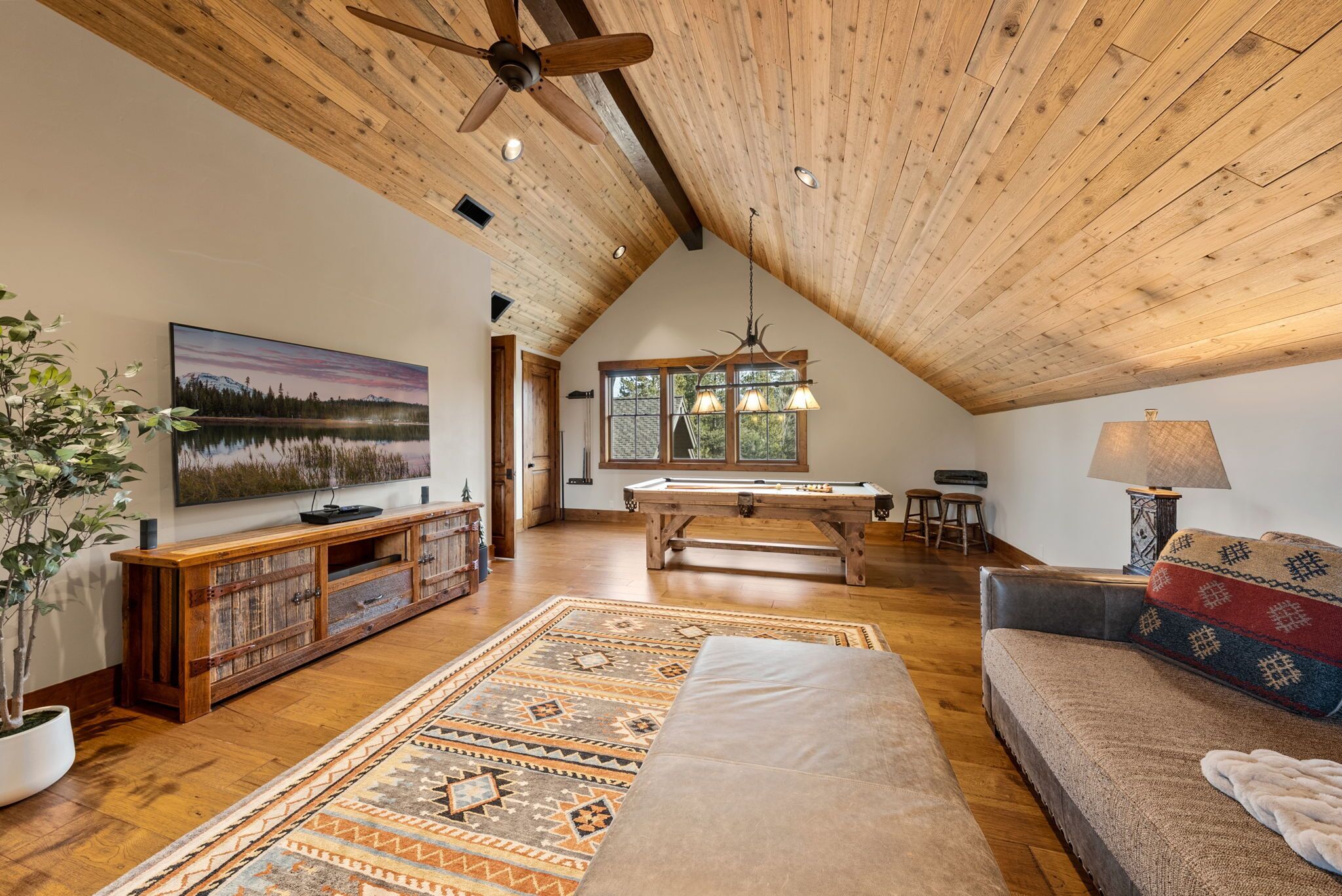 A spacious loft with a high ceiling, wooden beams, a pool table, a sofa, a TV, and a large window offering a view of the outdoors.